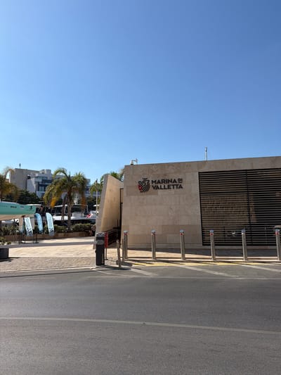 The image depicts the entrance to Marina di Valletta in Floriana, Malta. The main structure is a modern building with a beige stone facade, featuring the "MARINA DI VALLETTA" logo and a distinctive latticed metal section on the right. In front of the building, there is a barrier gate with bollards. To the left of the building, a section of a light-green boat with "BENETEAU" branding is visible, along with several promotional flags. Palm trees and other greenery are present, and in the background, there are modern residential or office buildings and several boats docked. The scene is set on a clear, sunny day, with a bright blue sky overhead. The foreground shows a paved area and a street. There are no people clearly identifiable in the image engaging in any specific actions.