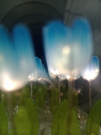 The image displays a close-up, slightly out-of-focus view of an illuminated artificial floral arrangement. Multiple objects resembling tulips are visible, featuring translucent plastic petals that transition from a vibrant blue at the top to a milky white with an internal glow towards the base. Each flower is supported by a twisted metallic wire stem. Below the petals, distinct, elongated green leaf-like structures are present, embedded in a clear, crystalline or granular medium that reflects light. The background is dark and indistinct, showing a curved, shadowy architectural element, possibly an archway or concave wall feature, which recedes into darkness. No individuals are present in the scene. This arrangement is located in Lomas de Zamora, Argentina.