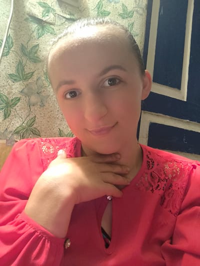 A young woman with light skin, dark eyes, and light brown hair tied back is shown in a close-up selfie. She is wearing a bright pink blouse with lace detailing around the neckline and cuffs. Her right hand is resting on her chin and neck. She is smiling slightly. The background is a patterned wallpaper with green leaves and white flowers, and a dark blue wall with white trim. The lighting suggests an indoor setting, possibly during the day. The overall mood is casual and friendly. There is no visible text in the image.