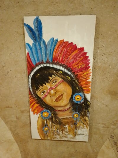 A vertical, full-body painting of a young Indigenous girl wearing a feathered headdress adorns a beige, textured wall in Cuiabá, Brazil. The girl has dark hair, brown eyes, and is adorned with face paint and a necklace. The headdress features blue and red feathers, and she wears blue flowers in her hair. The background of the painting is white, and the artist's signature is visible at the bottom right. The wall appears to be part of an outdoor or semi-outdoor space, possibly a courtyard or alleyway, with tiled flooring visible in the lower right corner.
