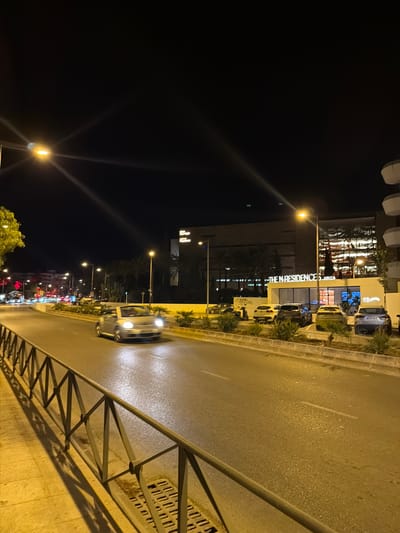 An exterior night-time street-level shot captures a street scene in Ibiza, Spain, featuring several cars, a building with illuminated signage, streetlights, and a sidewalk. The street is wet and reflective under the illumination of streetlights, with a silver convertible car in the foreground on the right, heading away from the camera. A dark metal fence with a zigzag pattern runs along the sidewalk on the left, casting long shadows. In the background, across the street, is a building with the illuminated text "THE N RESIDENCES IBIZA". Several parked cars are visible in front of this building. The sky is dark, indicating nighttime, with a few light sources creating streaks of light due to lens flare.