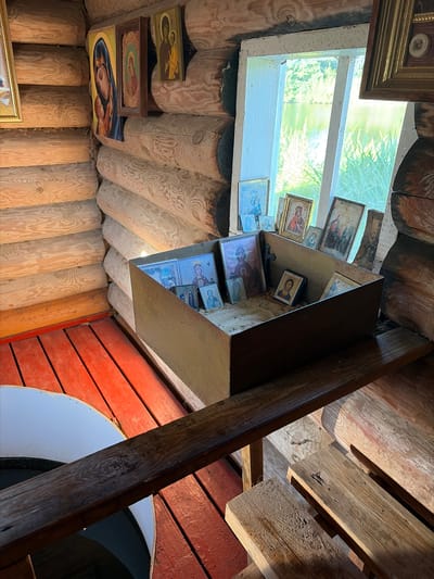 The image shows the interior of a log cabin, likely a small chapel or prayer space, located in Pobedinka, Russia. The walls are constructed from rough-hewn logs, and the floor is made of red wooden planks. A window with white trim looks out onto greenery and water, suggesting a natural setting.
In the center of the image is a metal box filled with sand, containing numerous small religious icons. These icons depict various saints and religious figures, some framed and others unframed. They are arranged in a seemingly informal manner, with some leaning against each other and others lying flat in the sand.
To the left of the box, on the log wall, are several larger icons. One prominent icon on the far left is a depiction of Mary and Jesus, with a dark blue veil. Next to it is another framed icon showing a similar religious scene. To the right of the central box, more icons are visible, some leaning on the windowsill.
In the foreground, a wooden beam extends horizontally across the lower portion of the image, with what appears to be a circular opening covered by a white surface to the left. To the right of the beam, wooden benches are visible. The overall impression is one of a humble, rustic spiritual space. There are no people visible in the image.