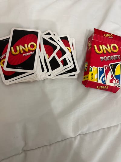 A red and yellow "UNO Pocket" card game box is partially opened, revealing a glimpse of the game cards inside. A stack of "UNO Pocket" cards, with red and black card backs, sits below the box on a white quilted surface. The "UNO Pocket" logo is prominently displayed on the card backs. Small print indicating "© 2014 Mattel" is visible on some cards.