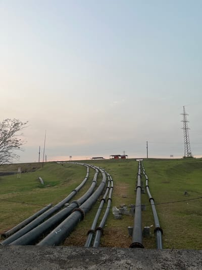 The image depicts an industrial landscape in Besuki, Indonesia, characterized by a series of large, dark pipes laid across a grassy embankment. The pipes, some appearing to be made of metal and others of a synthetic material, are arranged in parallel lines and curve upwards along the slope. They are supported by concrete structures and metal stands, indicating their function in transporting fluids, likely for industrial or water management purposes.

In the background, a tall, lattice-like electrical pylon dominates the skyline, suggesting the presence of power infrastructure. Several small, red-roofed buildings are visible near the crest of the embankment, possibly associated with the industrial operation. A lone tree with sparse foliage stands on the left side of the frame, while the sky is overcast with subtle hues of pink and orange, suggesting it might be dawn or dusk. The foreground is occupied by a rough, concrete surface, hinting at a man-made structure or pathway. No people are visible in the scene. The overall impression is one of functional infrastructure within a natural, albeit somewhat barren, environment.