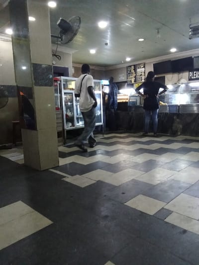 The image shows a black and white tiled floor in a food establishment in Ovwian, Nigeria, located at 5.503 latitude and 5.799 longitude. Three people are present, two men and one woman. The two men are both wearing casual clothing, with one of them walking towards the left of the image and the other standing in the center of the image facing left. The woman is wearing a black top and blue jeans and is standing near the counter, facing left, hands on her hips. The counter contains glass-fronted display cases with food items inside. In the background, a ceiling fan can be seen near the top left corner of the image. The walls are made of plain concrete and are painted in a light gray color. The room is well lit with fluorescent lights.
