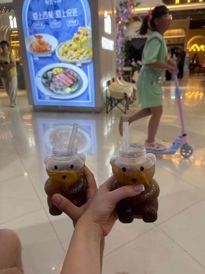 In Harbin, China, a person's hands are holding two bear-shaped beverage cups. The drinks are dark brown with a straw in each. In the background, a young girl with a pink bow in her hair and wearing a light green outfit rides a blue and pink scooter. To the left, a woman wearing a yellow shirt and beige pants stands near a brightly lit blue advertisement featuring images of food, including spaghetti, pizza, and steak. The scene appears to be indoors, possibly in a shopping mall or entertainment venue, with a reflective floor and decorative lighting.