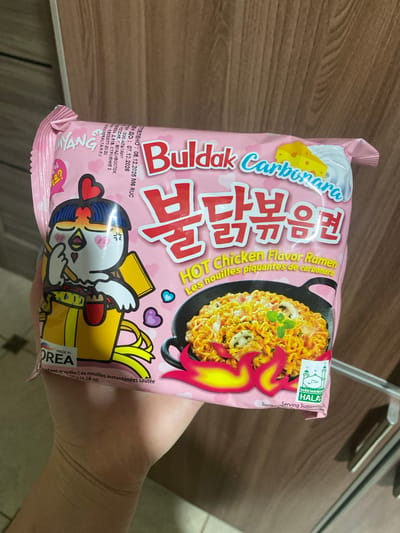 A person's hand holds a pink package of Buldak Carbonara instant noodles, showcasing the product against a brown wooden surface. The package is the central subject, featuring vibrant graphics.

The instant noodle packaging is predominantly pink and advertises "Buldak Carbonara HOT Chicken Flavor Ramen" in English and "불닭볶음면" in Korean. French text, "Les nouilles piquantes de carbonara" and "Les nouilles instantanées sautées," is also visible. A cartoon chicken mascot with blue hair and a heart clip is depicted on the front, along with an image of cooked ramen noodles in a black skillet, adorned with mushrooms and a chili pepper graphic in fiery red and yellow. A white cloud-like shape representing cream or sauce, and a yellow block of cheese, are also part of the design.

A green "HALAL" certification logo is visible in the top right corner. The package indicates "MADE IN KOREA" (partially obscured) and a weight of "130g (4.58 oz)." Russian text provides manufacturing and expiration dates: "ИЗГОТОВЛЕНО : 08.12.2025" (Manufactured: December 8, 2025) and "ГОДЕН ДО : 07.12.2026" (Best before: December 7, 2026). It also details the Russian importer, "ООО АЛЬТАИР" in Vladivostok, Russia, including an address and website "WWW.MIRALTAIR.RU". The background features a light brown wooden surface, possibly a table or counter, and a metal rack or handle in the upper right corner.