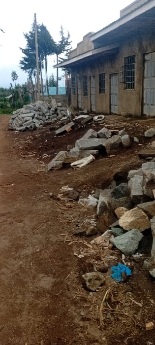 A building with barred windows sits beside piles of gray stones and construction debris. The ground is dirt, scattered with straw and a blue cloth. A stack of rectangular blocks rests at the edge of the frame, near a tree and some greenery. The photograph is taken at an angle, possibly with a wide-angle lens, resulting in some distortion.