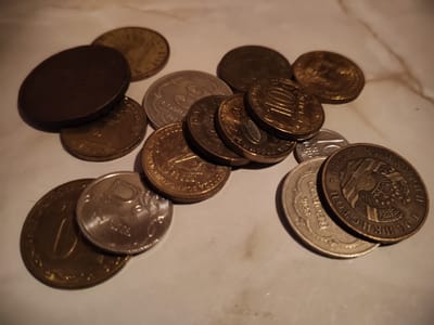 The image displays a collection of assorted coins scattered on a marble-like surface. The coins vary in size, color, and design, suggesting they are from different origins or denominations. Some coins are gold-colored, while others are silver-colored or appear to be made of a darker metal. Several coins have visible inscriptions and images, such as numbers, symbols, and portraits. One coin in the foreground, a silver-colored one, has a prominent "P" symbol. Another gold-colored coin shows a depiction of a bird or eagle. The lighting in the image creates shadows, indicating a single light source. The background is a light-colored, possibly tiled or marbled, surface. There are no people or animals present in the scene. The image is a close-up shot, focusing entirely on the coins and the surface they rest on. The location of Венюково, Russia is not ascertainable from the image.