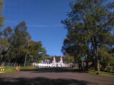 The image displays an outdoor scene in Batulawang, Indonesia, centered on a distinctive architectural complex viewed from a pathway. A dark, unpaved or dirt path dominates the lower central foreground, flanked by grassy areas. On the left, several portable barriers, horizontally striped orange and white, are positioned on the grass, with a chain-link fence visible behind them. Trees with green foliage line both sides of the path, casting shadows onto the ground, and extending into the midground. In the background, a light-colored building complex with multiple arches and two prominent, multi-tiered towers topped with reddish-pink pointed roofs is centrally featured. The building exhibits a decorative or whimsical architectural style. Hilly terrain covered in trees rises behind the structure. The sky is clear and deep blue, indicative of bright daylight. No individuals or specific activities are present within the frame.