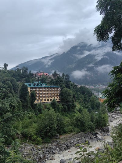 This image depicts a landscape in Aleo, India, characterized by a hotel situated on a verdant hillside overlooking a rocky river. The hotel, a prominent structure with a yellow facade and a green roof, features numerous balconies. It is nestled amongst dense evergreen trees. In the background, mist-shrouded mountains rise, their slopes covered in trees. The river, appearing somewhat turbulent and murky, flows over a bed of stones and rocks. The sky is overcast, contributing to a subdued atmosphere. Several other buildings are visible on the slopes and in the distance, suggesting a populated area. There are no identifiable people or specific actions occurring within the frame.
