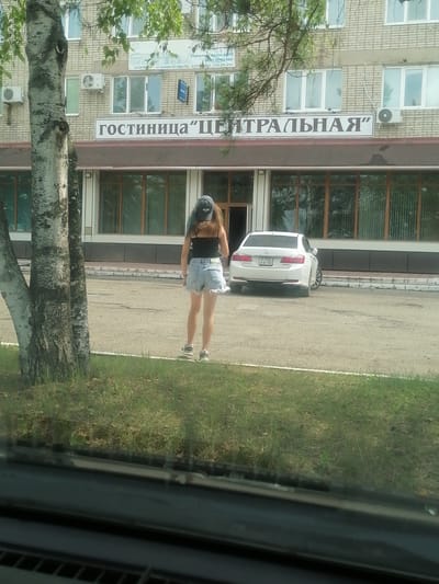 In Lesozavodsk, Russia, a young woman stands facing away from the camera, her back to a building with a sign that reads "ГОСТИНИЦА "ЦЕНТРАЛЬНАЯ"", which translates to "HOTEL 'CENTRAL'". She is wearing a black tank top, denim shorts, a dark baseball cap, and sneakers. A white car, a Honda Accord with license plate P 310 XK 125, is parked in front of the hotel. The scene is set on a paved area, with grass and trees visible in the foreground and background. The building appears to be a multi-story structure with numerous windows. The time of day is indeterminate due to the overcast lighting.