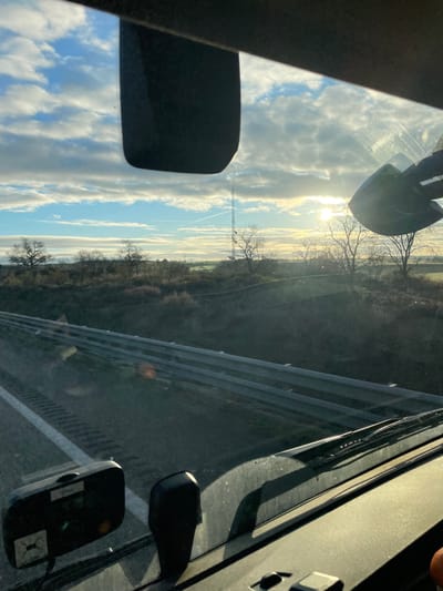 The scene depicts a view from the interior of a vehicle, looking forward through the windshield onto a multi-lane asphalt road and a rural landscape located near Fraga, Spain. The image captures a sky with scattered stratocumulus clouds and a low sun on the right horizon, causing lens flare and glare. A distinct white contrail from an aircraft is visible in the upper left portion of the sky.

In the foreground, the dark dashboard of the vehicle is visible, along with the base of the windshield where dark wiper blades rest horizontally. A rectangular, dark electronic device, possibly a toll transponder or dashcam, with a green illuminated indicator and a yellow dot, is mounted on the lower left area of the windshield. A portion of a black rearview mirror is visible at the top center of the frame.

To the left, the dark asphalt road features multiple parallel white dashed lane markings. A silver-colored metal guardrail separates the road from an uncultivated embankment below, which is covered with dry, sparse vegetation. Beyond this, the landscape extends into gently rolling terrain dotted with numerous deciduous trees, which are currently bare of leaves, indicating a cold season. A tall, open-lattice telecommunications tower stands prominently in the mid-ground, slightly to the right of center, against the brightening sky. No individuals or specific actions beyond implied vehicle travel are depicted.