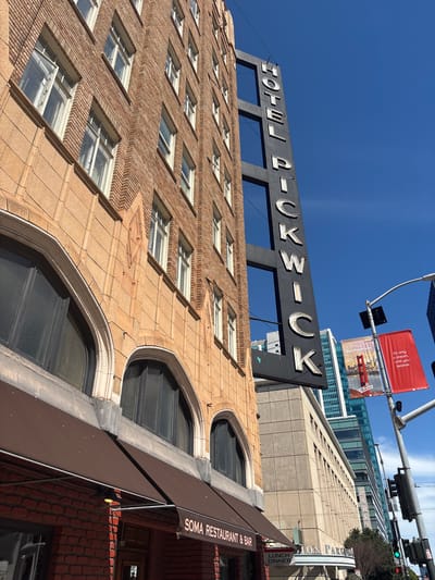 The image shows a low-angle exterior view of the Hotel Pickwick building in San Francisco, California, United States of America. The building is constructed of light brown brick with decorative stone accents around arched windows. A large, vertical black sign with illuminated white letters spelling "HOTEL PICKWICK" is attached to the side of the building. In the foreground, a brown awning covers the entrance to "SOMA RESTAURANT & BAR." Beyond the Hotel Pickwick, modern glass skyscrapers are visible against a clear blue sky. Traffic lights and banners are present on the right side of the image.