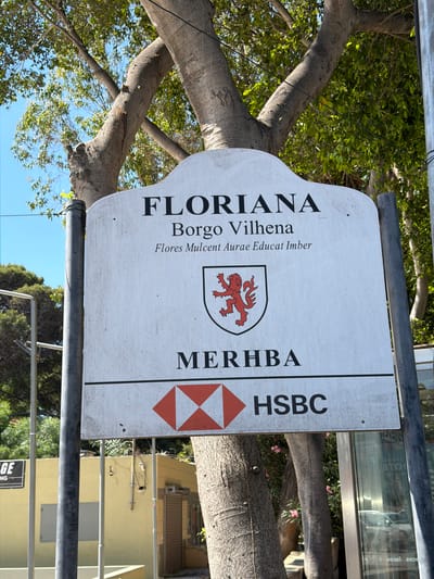 This is a medium shot, taken at eye level, of a sign in Pietà, Malta. The sign is white with a curved top and reads "FLORIANA Borgo Vilhena Flores Mulcent Aurae Educat Imber" in black text. Below this text is a shield with a red lion rampant. Further down, the sign reads "MERHBA" and displays the HSBC logo. The sign is supported by dark grey poles and is partially obscured by the trunk and branches of a large tree. In the background, a yellow building with a rolling shutter door is visible, as is a glass-fronted building with reflections of cars. The sky is clear and blue.
