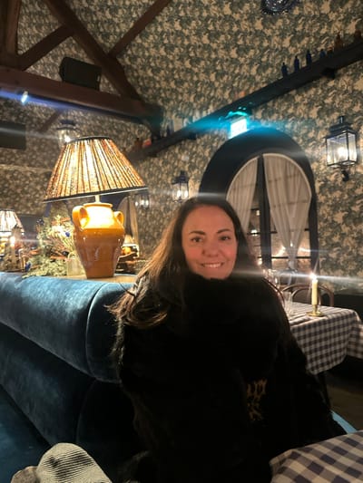 A woman smiles at the camera, seated on a plush, dark blue couch. She wears a black top with a leopard print under layer. Overhead, there's a decorative window, framed by white curtains. A lantern-style light fixture hangs nearby. A lamp with a patterned shade sits close, illuminating a pottery vase. The room's decor features a leafy wallpaper, exposed wooden beams, and holiday decorations visible in the background.