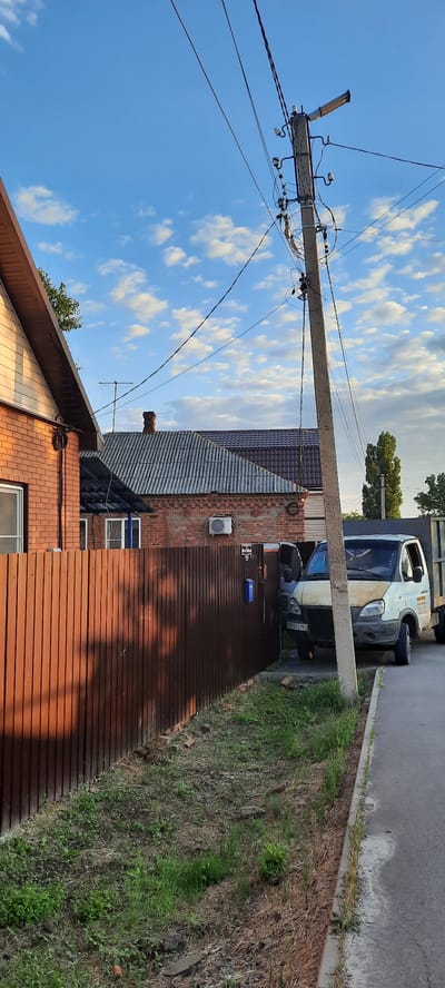 The image depicts a street scene in Shakhty City District, Russia, under a cloudy blue sky. In the foreground, a brown corrugated metal fence runs along the left side of the frame. Beyond the fence, a brick house with a light-colored siding and a dark roof is visible. Further back, more houses with brick walls and corrugated roofs are seen, along with a chimney.
Dominating the center of the image is a concrete utility pole adorned with numerous wires and insulators. A street light is attached to the top of the pole. To the right of the pole, a white panel van with a yellow hood is parked, facing forward. The van has a license plate that appears to read "A470TC 161". Another vehicle, a dark truck with a gray cargo bed, is partially visible behind the van. The ground beside the road is covered in sparse grass and dry vegetation. The overall impression is of a quiet residential street with overhead power lines.