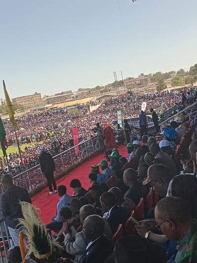 This image captures a large outdoor public gathering or rally, likely held in Siaya, Kenya, viewed from an elevated perspective. The scene is filled with a vast crowd of people extending into the distance under a clear, bright sky, indicating daytime with sunny weather.
In the foreground, individuals are visible from above, many seated in red stadium-style seats. Their heads and backs are mostly visible, with some appearing to look forward or downwards, engrossed in the event. A broad red carpet or covered area forms a prominent path or stage extending from the immediate foreground into the mid-ground.
The mid-ground is densely packed with attendees, forming a sea of people. Interspersed within the crowd are several banners and flags. A red banner with white text is clearly visible, displaying "LUO" and the year "2013". Other banners, including one with green and white colors, are also present but their text is illegible. Two tall, slender poles or masts rise above the crowd in the mid-distance.
The background reveals a flat, open landscape with some distant buildings, consistent with an urban or semi-urban environment. A faint, thin line, possibly from a kite or a distant aerial object, is visible against the clear sky in the upper left. The overall impression is one of a significant, well-attended public assembly.