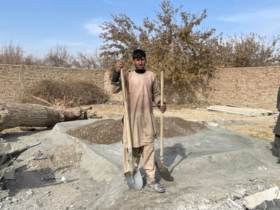 This image depicts an outdoor construction or clearing site in Nakhonay, Afghanistan. Centered in the foreground is an adult male, dressed in a light tan long-sleeved tunic over a dark undershirt and light trousers, with open-toed sandals. He looks directly at the camera, holding a long-handled shovel vertically in his right hand and a shorter-handled shovel angled downwards in his left hand. To his immediate right, the lower body of another individual in dark clothing is partially visible. Behind the central figure, a pile of gravel and sand is partially contained by an irregular, low concrete curb with sloped sides. To the left, a large, felled tree trunk lies horizontally near a partially visible metal wheelbarrow. Further back, a substantial mound of dry brush or woody debris is positioned against a light-colored brick wall that spans the mid-ground. The background beyond the wall features several leafless deciduous trees under a clear, light blue sky. The ground throughout the scene is uneven, light-colored soil with scattered rocks and construction debris. Shadows suggest strong sunlight from the upper right.