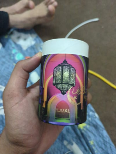 An overhead, medium shot shows a hand holding a cylindrical container with a white top. The container has a colorful, patterned label that features a black lantern, a gold crescent moon, a star, and Arabic calligraphy. The text "AJMAL" is visible in the lower center of the label. The background is blurred and shows a blue patterned fabric, brown flooring, and out-of-focus feet and legs. There is no indication of the specific location in Abbottabad, Pakistan, or any context that would suggest this city.