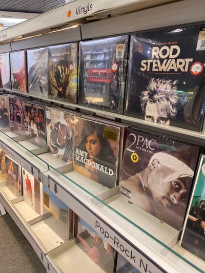 This image captures a section of a retail store, likely a music or department store, showcasing an array of vinyl records displayed on shelves. The setting is indoors, with records neatly arranged vertically in clear protective sleeves within metal and glass display cases.
The main subjects are the numerous vinyl albums. Prominently featured on the left side, from top to bottom, are "THE MANY FACES OF ROD STEWART" and "THE MANY FACES OF RAMONES." To their right, higher up, is a record by "2PAC" titled "LIVE MY LIFE," which also carries a "PARENTAL ADVISORY EXPLICIT CONTENT" label. Below the 2Pac album is an album by "AMY MACDONALD" titled "THIS IS THE LIFE." Other album covers are partially visible further down and to the right, but their details are less clear. Many of the records have price or information stickers, with some indicating "180g" (presumably for heavy-weight vinyl) and QR codes.
The activity implied is shopping for music. The shelving system uses clear glass separators and metal frames. On the far left, a white sign reads "Vinyls" with a French translation "Disques" below it, hinting at a French-speaking location, consistent with Liège, Belgium. Another sign to the right categorizes records as "Pop-Rock K-L" and further down, "Pop-Rock N-." The overall impression is that of a well-organized music section in a store. No people are visible, and the time of day is undeterminable.
