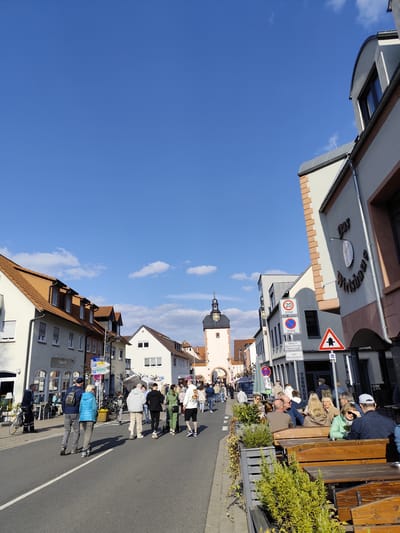 The image depicts a daytime street scene in Obernburg am Main, Germany, looking east towards the Obertor (Upper Gate). A clear blue sky with scattered white cumulus clouds is visible overhead. The asphalt street, marked with a white dashed centerline, is occupied by approximately two dozen pedestrians walking in various directions, with a concentration moving towards the gate in the background.
On the left side of the street, a sequence of multi-story buildings with light-colored facades and terracotta-tiled pitched roofs includes ground-floor businesses; a silver bicycle is parked upright near the curb in front of one.
On the right side, a prominent building identified by the sign "Das Wirtshaus" features a grey and peach-toned facade with a distinctive curved roofline. An outdoor dining area associated with this establishment contains numerous wooden tables and benches, occupied by approximately 15 individuals engaged in seated activity. This area is delineated from the roadway by planters containing green shrubbery. Adjacent to "Das Wirtshaus" are multiple German traffic signs, including two circular signs indicating a "20 ZONE" speed limit, a triangular warning sign for a priority road, and a rectangular sign detailing paid parking ("Parken ist gebührenpflichtig mit Parkschn").
The Obertor, a historic clock tower constructed of light-colored stone with a dark, conical roof and spire, stands centrally in the background, featuring an arched passage at its base. All individuals depicted are dressed in casual attire. Natural sunlight illuminates the scene, creating distinct shadows.