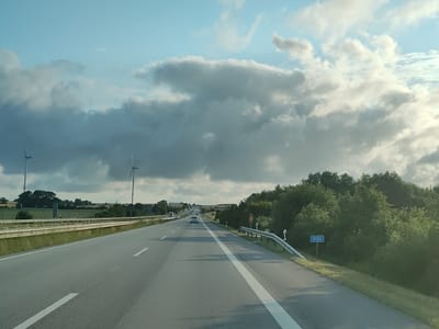 The image shows a highway in Gremersdorf, Germany. The highway is paved and has two lanes in each direction. There are guardrails on both sides of the road. On the left side of the road, there is a field with some trees and two wind turbines in the distance. On the right side of the road, there is a dense forest. The sky is cloudy and overcast, with some patches of blue sky visible. There is a sign on the right side of the road that reads "117.5". There are no people or vehicles visible in the image, except for a white car in the distance on the highway. The image appears to have been taken from a moving vehicle.