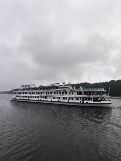 A large white river cruise ship, named "VOLGA DREAM," sails on dark, choppy water under a cloudy sky. The ship has multiple decks, with rows of windows and balconies. A Russian flag is visible on the stern. Lush green trees line the distant shore. The ship is moving from left to right in the frame, leaving a wake behind it.