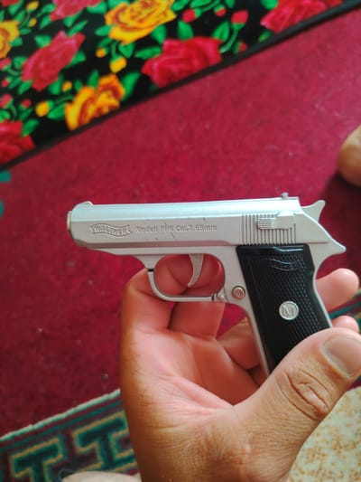 A silver toy pistol is being held by a person's hand. The pistol has "WALTHER Modell PPK Cal. 7.65mm" inscribed on its slide. The background consists of a red carpet with a floral pattern and a rug with a geometric design. The image appears to have been taken indoors, possibly in a residential setting in Khujand, Tajikistan.