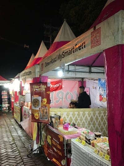 A food stall is visible, with a pink and white awning and banners advertising “MySmartfren” and “TIBA-TIBA WONTON." The wonton sign highlights a price of 10 or 15, presumably in local currency. A person in a black hoodie is seen inside the stall, possibly serving customers. Various food products are displayed on the table. A tiled surface suggests this is a sidewalk location. The environment is dimly lit, indicating it is likely evening or night.