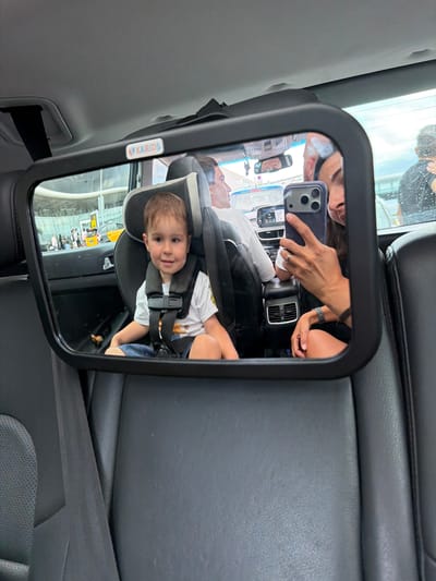 This image captures a moment inside a vehicle, likely a taxi, as indicated by the presence of a taxi in the reflection of the mirror and the overall context of travel. The primary focus is a young boy, approximately 3-4 years old, seated in a car seat in the back of the vehicle. He is wearing a white t-shirt with a graphic print and blue shorts, secured by a car seat harness.

A wide-angle mirror, mounted on the headrest of the seat in front of the boy, reflects the interior of the car and its occupants. In the mirror, we see a woman holding a smartphone, taking a selfie. She is wearing a dark top and has a ring on her left hand. To her left, a man is visible in the reflection, looking forward. The background seen through the car's windows and reflected in the mirror suggests an airport environment, with a modern terminal building and a yellow taxi visible outside. This indicates the location is likely El Prat de Llobregat, Spain, specifically at or near Josep Tarradellas Barcelona–El Prat Airport. The car's interior appears to be upholstered in dark leather, with visible air vents and a dashboard. The overall scene suggests a family or group traveling, possibly to or from the airport.