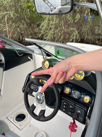A view from inside a boat shows a hand gesturing with the index and middle fingers extended towards the steering wheel. The boat is parked near a wooded area. The location is not discernible from the image alone.
