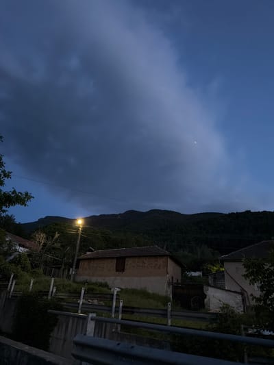 The image depicts a nighttime scene in Sapareva Banya, Bulgaria. In the foreground, there is a metallic guardrail, and beyond it, a building with a tile roof and concrete walls. A street lamp illuminates the building and a section of the nearby forest that ascends a mountain. The sky is overcast with heavy, dark clouds, and a sliver of a crescent moon is visible. Power lines stretch across the scene. The environment suggests a rural or semi-rural area on the outskirts of the town.