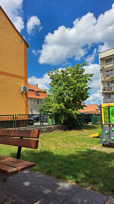 The image depicts a sunny day scene in Krosno Odrzańskie, Poland, with a focus on a residential courtyard or small park area. In the foreground, a wooden bench is positioned on a paved pathway made of dark gray interlocking stones. The pathway is partially shaded by the bench. Beyond the bench and pathway, a patchy, green lawn stretches out, interspersed with some dry brown grass.
To the left, a portion of a tall building with an orange-yellow stucco exterior dominates the left side of the frame. An outdoor air conditioning unit is mounted on its wall. Behind this building, a smaller residential building with a red-tiled roof is visible, featuring windows and a chimney. Adjacent to this building, a dark-colored car is parked behind a green metal fence.
In the center of the image, a large, leafy green tree stands prominently. The tree has numerous leaves and some lighter green or yellowish elements, possibly flowers or new growth. Behind the tree and further back, several large, dark green bins or dumpsters are lined up, also behind a green metal fence.
To the right of the tree and fence, a section of a colorful playground structure is visible. It is predominantly black and yellow, with green climbing grips or panels. A yellow slide extends from the structure.
In the background, a multi-story apartment building with a white facade and balconies can be seen. The sky above is a vibrant blue, dotted with fluffy white clouds, indicating fair weather. The overall scene suggests a residential area with common amenities like a playground and green spaces.