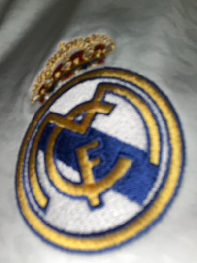 A close-up shows the embroidered logo of Real Madrid. The central design features interconnected letters outlined in gold, layered over a blue, vertical stripe on a white background, enclosed within a golden double-ringed circle. The crown is visible, embroidered in gold and shades of red, positioned to the left of the main emblem. The emblem appears on a gray fabric.