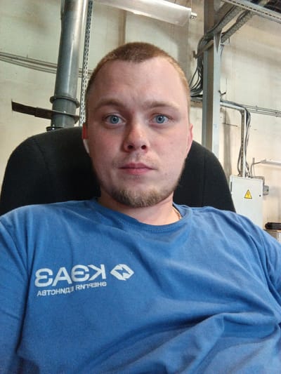 This media file shows a close-up, front-facing selfie of a young man with light skin, blue eyes, and short brown hair. He has a light beard and is wearing a blue t-shirt with a logo. The logo appears to be in a foreign alphabet, possibly Cyrillic, and includes a stylized symbol that resembles a recycling logo. The man is wearing white wireless earbuds.
He is seated in a black office chair. In the background, industrial elements are visible, including a large gray pipe, electrical conduits, and a light-colored wall. There is also a yellow triangular warning sign on a white electrical box. The overall setting appears to be an indoor industrial or workshop environment.
The lighting suggests it is daytime, but there are no clear indicators of the time of day or weather. The man's expression is neutral. The text on his shirt is not fully legible, but the prominent part appears to be "САЄК". Below this, there's another line of text that is partially visible and reversed, reading something like "АВТОНДЭ RN193HЭ". The man is in Kursk, Russia.