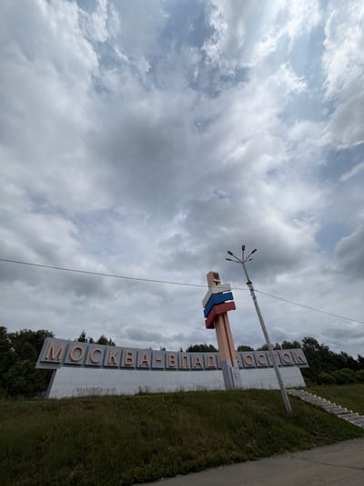 An eye-level, wide-angle shot captures a large, gray, angled sign with orange letters reading "МОСКВА-ВЛАДИВОСТОК" (Moscow-Vladivostok). The sign is situated on a grassy hill, with trees forming a dark backdrop behind it. To the right of the sign, a tall, weathered utility pole with multiple lamp fixtures extends upwards, leaning slightly to the right. A concrete staircase with several steps ascends the grassy hill to the right of the sign. In the foreground, a gray asphalt road runs along the bottom of the frame. The sky above is filled with dramatic, gray and white clouds. The scene appears to be an outdoor roadside landmark.