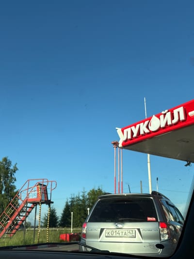 The image depicts a view from inside a vehicle at a gas station. The gas station's canopy, with the "Лукойл" (Lukoil) logo prominently displayed in white lettering on a red background, is visible in the upper right portion of the frame. The sky is a clear, bright blue.
To the left of the gas station canopy, a red metal staircase with a platform at the top stands out against the green grass and trees. This structure appears to be part of the gas station's infrastructure. In the middle ground, a silver Subaru Forester is parked, facing away from the viewer. The car has a license plate that reads "K014TA 43 RUS". Various stickers are affixed to the rear of the vehicle.
The environment suggests an outdoor setting, likely on a sunny day, given the clear sky and lighting. The presence of the gas station and the car indicates a typical scene at a service station. The location, Kirovo-Chepetsk, Russia, places this scene within a specific geographical context. There are no people clearly visible in the image. The overall impression is one of a routine stop at a roadside establishment.