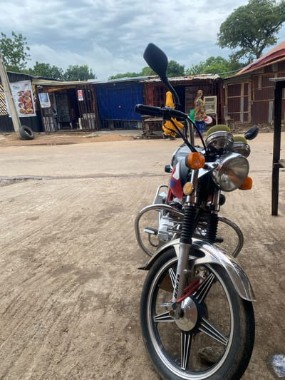 The image shows a motorcycle parked on a concrete surface in what appears to be an outdoor market or roadside stall area. The motorcycle is the primary subject, facing left and angled towards the viewer. It has a chrome finish, distinctive orange turn signals, and a yellow cloth draped over the handlebars. A red and white emblem is visible on the side of the motorcycle. The wheel features a multi-spoke design with the brand name "SUPER" visible.
Behind the motorcycle, a row of small shops or stalls with corrugated metal roofs and wooden or metal structures are lined up. One stall has a blue tarp or curtain. Several signs are visible, including one with "POS" and another displaying food items with the text "Choco". A person is partially visible near one of the stalls, wearing a colorful patterned garment and a white hat. Another person stands further back, near a window.
The ground is a worn, textured concrete, with some debris and a spare tire visible. Trees with green foliage can be seen in the background, suggesting a possibly rural or semi-urban setting. The sky is overcast with visible clouds, indicating a possibly humid or pre-rain weather condition. The overall impression is a snapshot of daily life in a West African marketplace, with the motorcycle serving as a common mode of transport or a vendor's possession. The scene conveys a sense of everyday commerce and activity.