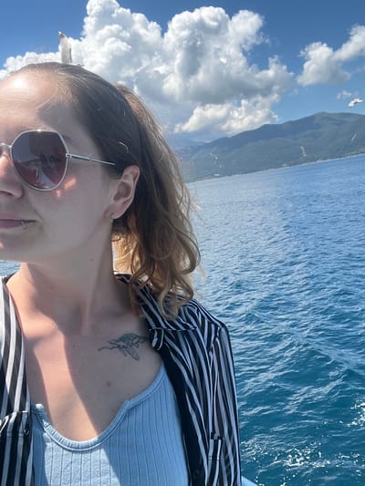 A woman is captured in a close-up selfie with a backdrop of the sea and mountains. She is wearing sunglasses and a striped shirt over a light blue top. A tattoo of a sea turtle is visible on her chest. The sea is a vibrant blue, with small waves. In the distance, a mountainous coastline is visible under a partly cloudy sky. Two seagulls are present, one perched on the woman's head and another flying in the distance. The image appears to be taken on a boat during the day. The presence of the sea and mountainous coastline suggests a location like Thassos, Greece, known for its natural beauty and proximity to the sea.