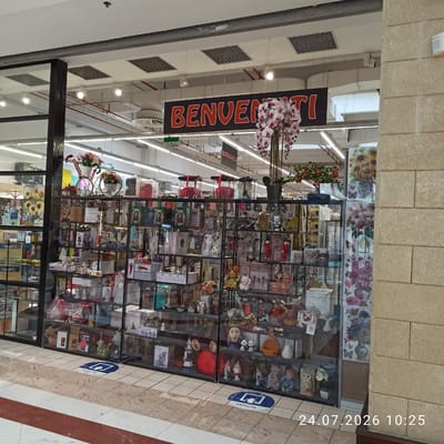 The image shows the storefront of a retail establishment in Modugno, Italy, featuring a display of various merchandise behind glass. A prominent banner above the entrance reads "BENVENUTI" (Welcome) in large red letters. The interior of the store is filled with shelves stocked with decorative items, giftware, and possibly electronics, indicated by a sign that says "ELETTRONICA". The displays include items like figurines, vases, artificial flowers, and packaged goods. The floor is tiled with a pattern of squares. The overall impression is that of a well-organized shop offering a range of consumer products. A date and time stamp at the bottom right corner indicates "24.07.2026 10:25".