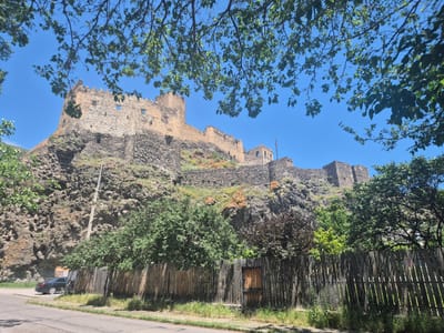 This media file captures a view of the Khertvisi Fortress in Georgia, a historic stone castle perched atop a rocky hill under a clear blue sky. The image is taken from a low angle, looking up towards the fortress, with the foreground dominated by a rustic wooden fence and some foliage from overhanging trees.

The fortress itself is the main subject, appearing weathered and ancient, with sections of crumbling stone walls and towers. The rocky terrain surrounding the fortress is visible, interspersed with patches of dry grass and some reddish-orange soil or rock. Green trees and shrubs grow at the base of the hill, partially obscuring the lower parts of the fortress and the fence.

In the foreground, a wooden fence runs horizontally across the image, with a weathered wooden gate in the center. To the left of the fence, a dark-colored car is parked on the side of a paved road. The road is visible in the lower left corner of the image. The grass and weeds growing along the roadside and at the base of the fence suggest a natural, somewhat unkempt setting.

The time of day appears to be daytime, with bright sunlight casting shadows, and the weather is clear and sunny, indicated by the vibrant blue sky and the overall lighting. The lush green leaves on the trees suggest a season other than winter, possibly spring or summer. The presence of the fortress, the rocky landscape, and the distant appearance of a utility pole hint at a rural or semi-rural historical location in Georgia. No people are visible in the frame. There is no discernible text in the image.
