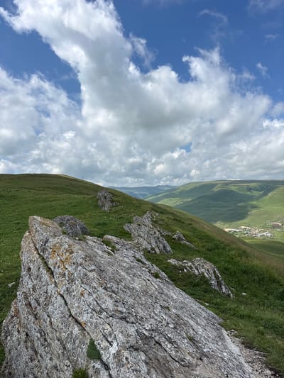 A vertical, medium shot shows a mountainous landscape under a partly cloudy sky. In the foreground, a large, weathered rock formation with lichen and moss sits on a grassy slope. The rock is textured and striated, with visible layers and cracks. Beyond the rock, a lush, green hillside slopes downwards, dotted with smaller rocks and patches of grass. In the middle ground, a series of rolling green hills recedes into the distance. A small village with houses and buildings can be seen nestled in a valley between the hills. The sky is a bright blue, filled with large, fluffy white clouds. The overall scene depicts a natural, undeveloped environment, likely in a rural area, showcasing the rugged beauty of the mountains and the tranquility of a remote village. This scene is located in Kichi-Balyk, Russia.