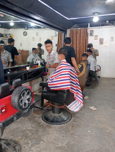 A barbershop interior in Pulorayeuk, Indonesia, is depicted with several people present. In the foreground, a child sits in a barber chair, draped in a red, white, and blue striped cape. To the left, a red toy jeep is partially visible, with a large tire and wheel. Behind the child, another barber chair is occupied by a person wearing a striped shirt, and a barber, wearing a green shirt, is attending to them. Further back, other individuals are seen, with a large mirror reflecting the barbershop's contents, including barber tools and equipment on a shelf. The walls are decorated with posters, and brown curtains hang in the background. The floor is concrete, and a ceiling with lights is visible overhead.