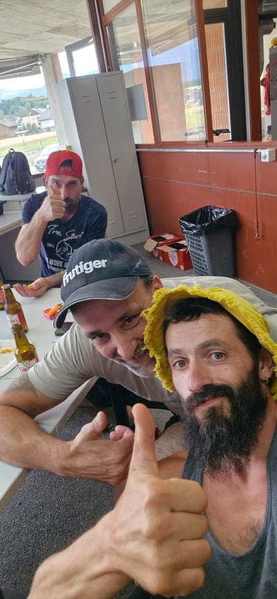 The image shows two men in an indoor setting, possibly an office or workshop. One man is wearing a grey t-shirt and a black baseball cap with "huttiger" written on it, and he is giving a thumbs-up gesture. The other man is wearing a blue t-shirt and a red baseball cap, and he is also giving a thumbs-up gesture. Both men appear to be happy and relaxed.
In the background, there are several objects, including a table with bottles of beer, a black backpack, a grey locker, and a trash can. The overall impression is that of a casual gathering or break time. The lighting suggests it is daytime. The image also includes a partial view of the exterior through a window, showing buildings and trees, hinting at an outdoor environment beyond the immediate setting.
There is text visible on the beer bottles and on the t-shirt of the man in the red cap. The t-shirt has white text that reads "Since 1919", "Work", and "Start". The bottles have text that appears to be in Portuguese.