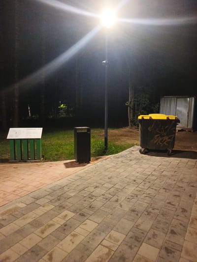 The image depicts a scene at night in a park or wooded area with a paved walkway. A bright streetlamp illuminates the scene, casting long shadows. To the left, there is a green-colored informational stand with a white sign. Adjacent to it is a dark-colored trash receptacle. Further to the right, a large, yellow-lidded dumpster with graffiti is visible. In the background, trees are silhouetted against the dark night sky, and a metal structure with a door stands to the far right. The paved walkway is made of interlocking stones, some of which appear wet. The location is identified as being in the city of Smolensk, Russia.