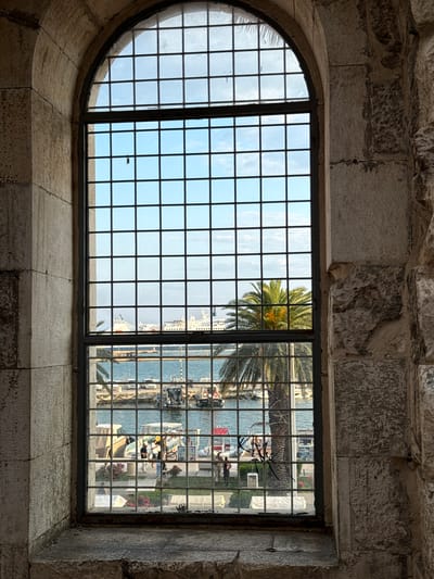 An arched window with a grid of dark metal bars looks out onto a harbor in Split, Croatia. The window is framed by weathered stone blocks. Through the window, a body of water with several boats is visible. In the distance, large white cruise ships are docked. A tall palm tree with green fronds stands prominently in the middle ground, partially obscuring the view of the ships. Several people are visible on a walkway near the boats, and parked cars are seen along the waterfront. The sky is blue with scattered clouds.