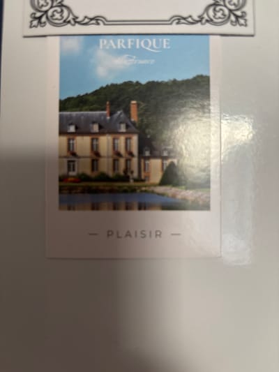 The image shows a postcard with a photograph of a French chateau or mansion. The chateau is a light yellow building with a dark grey roof and dormer windows. It is surrounded by trees and a body of water that reflects the building. Above the chateau, a blue sky with some clouds is visible. Text on the postcard reads "PARFIQUE de France" at the top, and "- PLAISIR -" at the bottom. The postcard is partially obscured by a white border and appears to be placed on a white surface. The overall impression is that of a travel souvenir or promotional item. There is no information in the image or its context to suggest any connection to the city of Imeni Morozova, Russia.