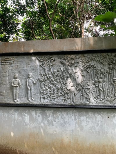 The image shows a stone relief sculpture depicting a group of people in a forest setting. The sculpture is mounted on a wall. The wall is made of concrete and has a light grey color. The sculpture is made of grey stone. The sculpture is framed with a black frame. The sculpture depicts a scene of a teacher teaching a group of students. The teacher is standing in front of a group of seated students. The teacher is holding a book and pointing at it. The students are listening intently and some are taking notes. The scene is set in a forest with trees in the background. The sculpture is located in Kecamatan Borobudur, Indonesia, at the coordinates -7.606, 110.181. There are no other people or objects present in the image.