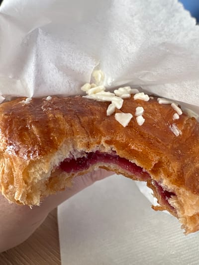 An overhead close-up shot captures a partially consumed pastry held by a human hand within white paper wrapping. The pastry exhibits a golden-brown, glossy exterior with visible laminated dough layers, characteristic of a Danish or croissant. A bite has been taken from its left side, exposing a bright red, jam-like filling. Irregularly shaped white sugar crystals or icing fragments are scattered across the top surface of the pastry. A light-skinned hand, visible on the lower left, supports the pastry. The white paper wrapping surrounds most of the pastry, and a light-colored, possibly wooden, surface is discernible in the lower-left background. The scene is located in Зеленогорское сельское поселение, Ukraine.